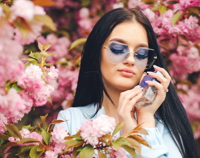 Sensual woman with perfume bottle in pink flowers. Spring pink sakura blossom. Spring day. Fashionable girl in trendy glasses